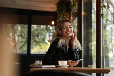 Photo of Young woman with headphones and smartphone listening to music in cafe