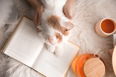 Woman with cute fluffy cat, tea and book on faux fur, top view
