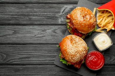 Photo of Tasty burgers, French fries with sauces on black wooden table, flat lay and space for text. Fast food