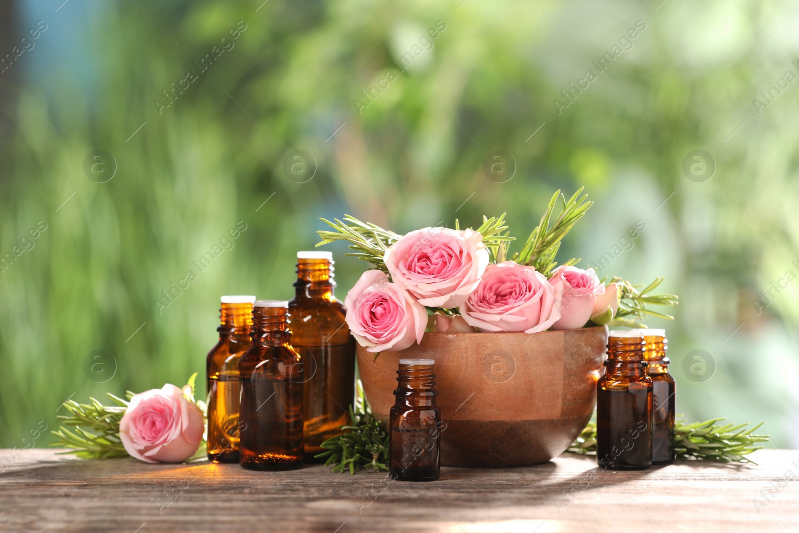 Photo of Bottles with essential oils, roses and rosemary on wooden table against blurred green background. Space for text