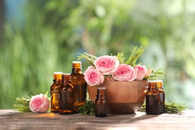 Bottles with essential oils, roses and rosemary on wooden table against blurred green background. Space for text