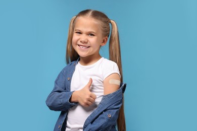 Photo of Happy girl with sticking plaster on arm after vaccination showing thumbs up against light blue background
