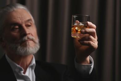 Photo of Senior man in suit holding glass of whiskey with ice cubes on brown background, selective focus