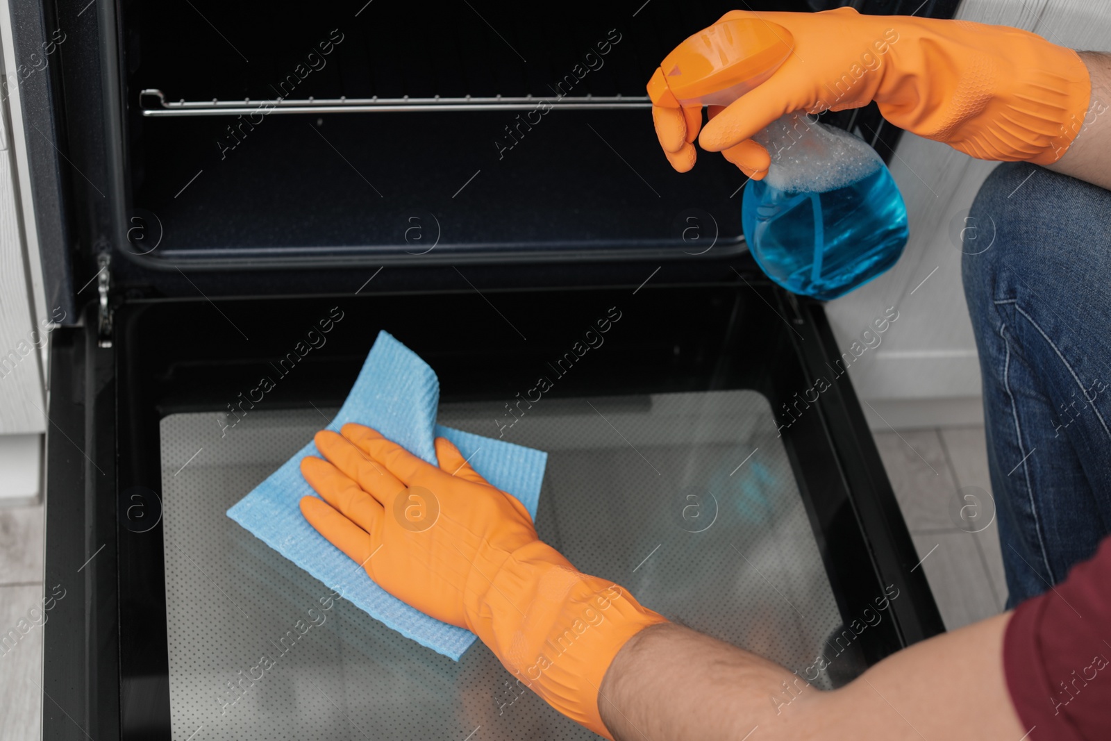 Photo of Young man cleaning oven with rag and detergent in kitchen, closeup