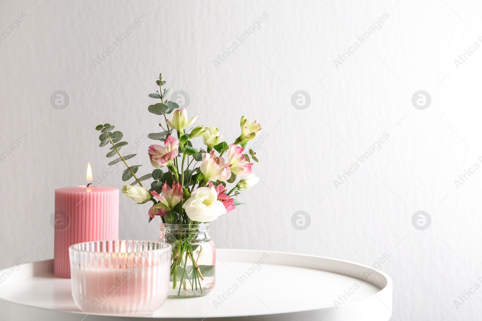 Photo of Stylish tender composition with burning candles and flowers on white table against light background, space for text. Cozy interior element