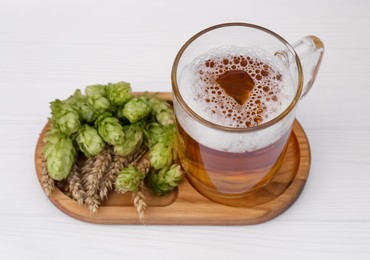 Photo of Mug with beer, fresh hops and ears of wheat on white wooden table, closeup