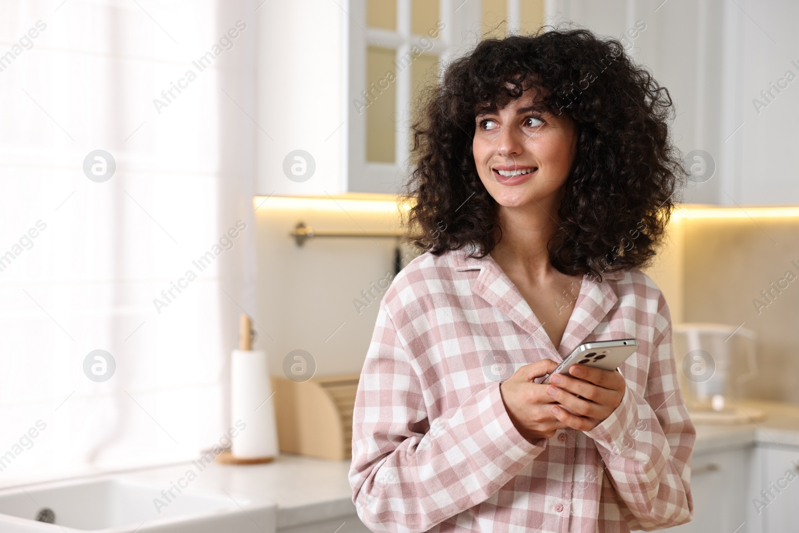 Photo of Beautiful young woman in stylish pyjama with smartphone in kitchen, space for text