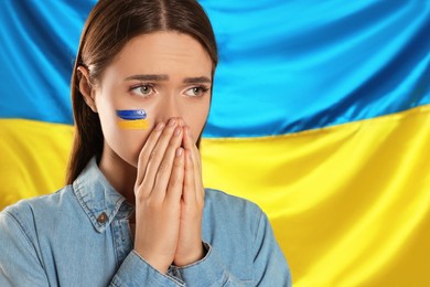 Sad young woman with clasped hands near Ukrainian flag. Space for text