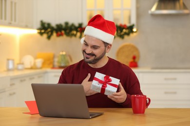 Celebrating Christmas online with exchanged by mail presents. Happy man in Santa hat thanking for gift during video call on laptop at home