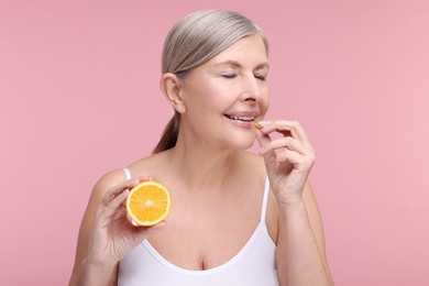 Photo of Beautiful woman with half of orange taking vitamin capsule on pink background