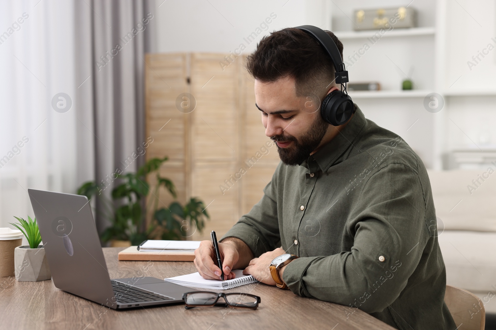 Photo of E-learning. Young man taking notes during online lesson at wooden table indoors