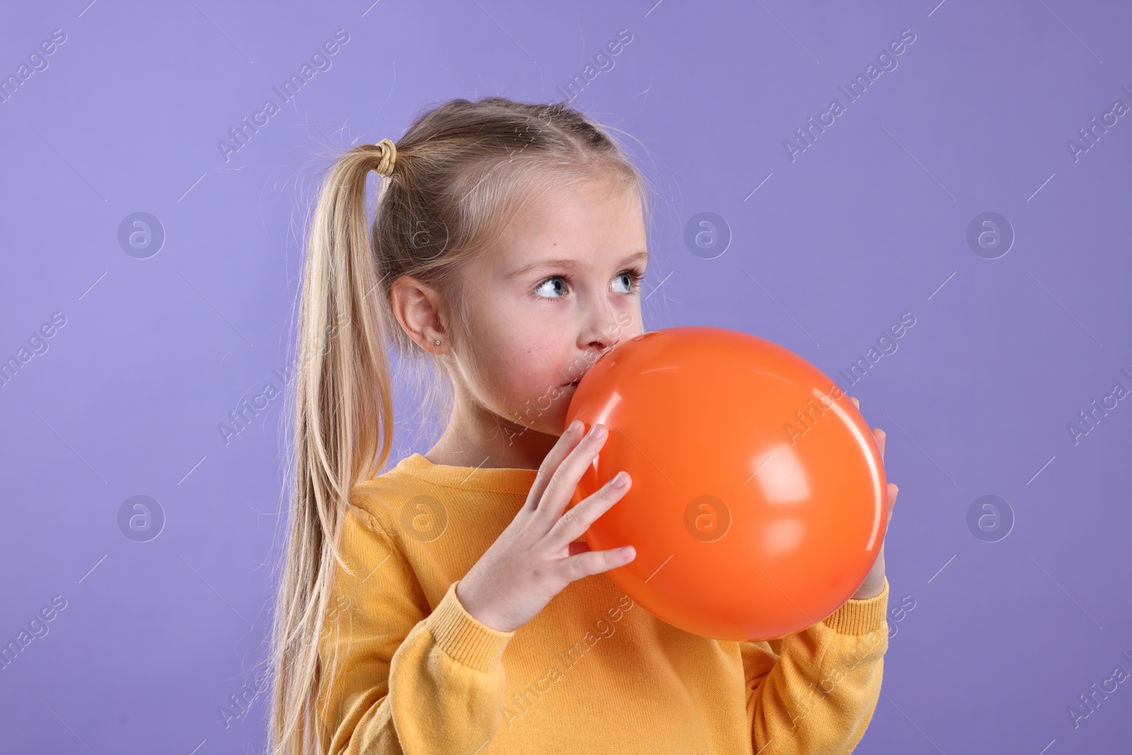 Photo of Cute little girl inflating orange balloon on violet background