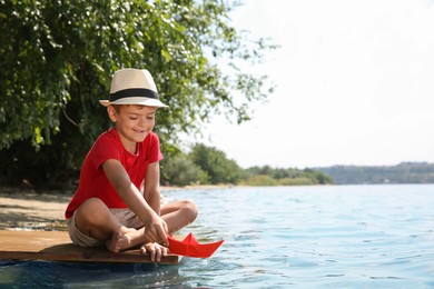 Photo of Cute little boy playing with paper boat on wooden pier near river