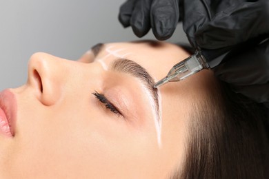 Photo of Beautician making permanent eyebrow makeup to young woman on grey background, closeup