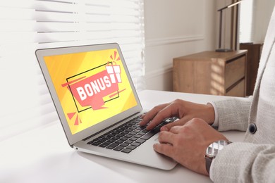 Bonus gaining. Man using laptop at white table indoors, closeup. Illustration of gift box and word on device screen