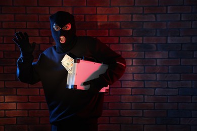 Photo of Emotional thief in balaclava with briefcase of money in color lights against red brick wall. Space for text
