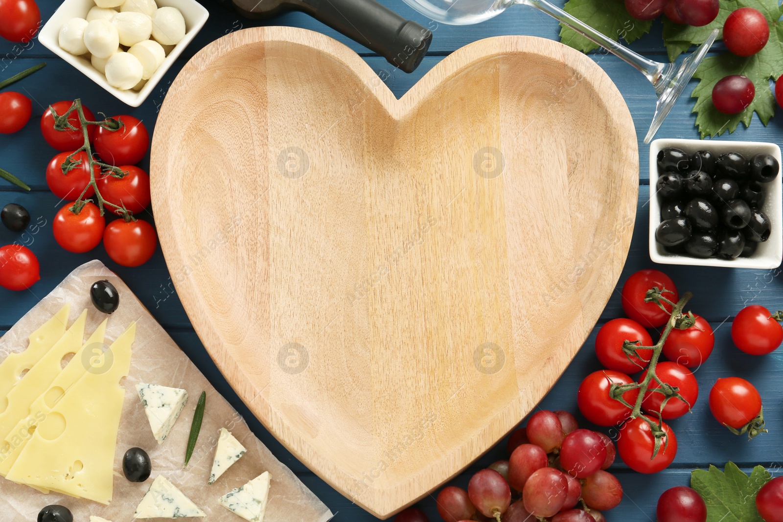 Photo of Heart shaped bowl, tasty red wine and snacks on blue wooden table, flat lay