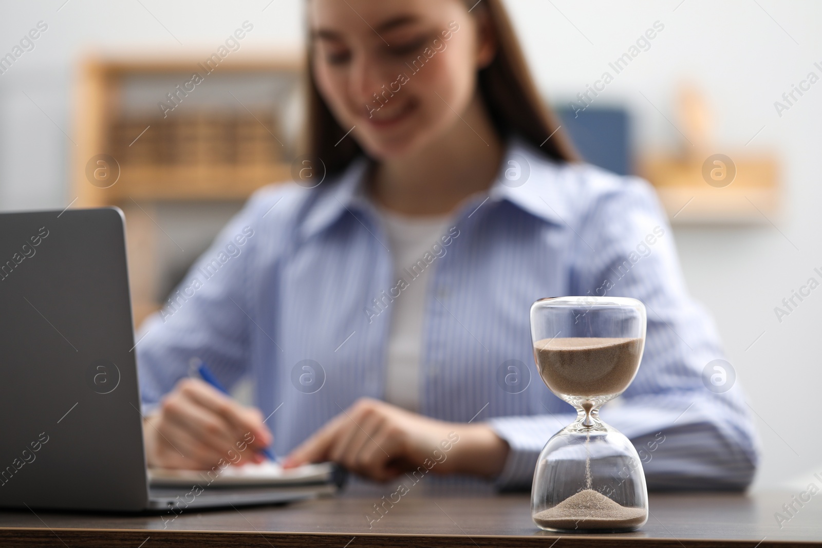 Photo of Hourglass with flowing sand on desk. Woman taking notes while using laptop indoors, selective focus