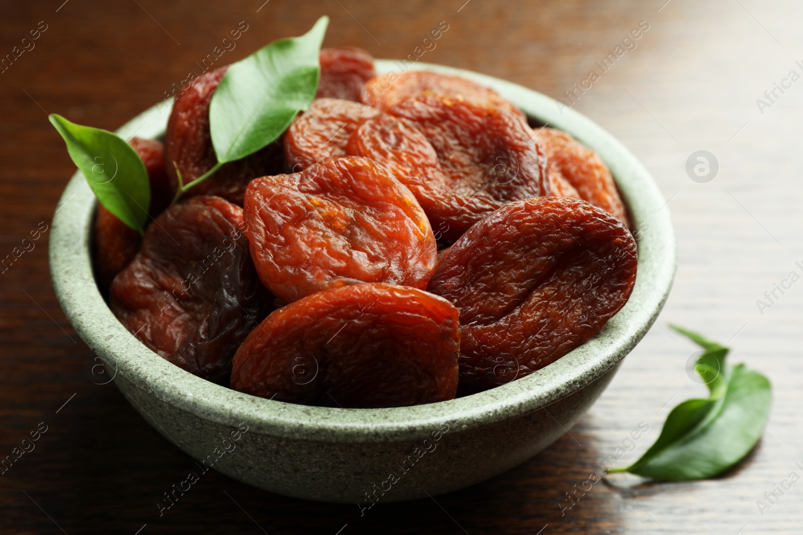 Photo of Bowl of tasty apricots and green leaves on wooden table, closeup. Dried fruits