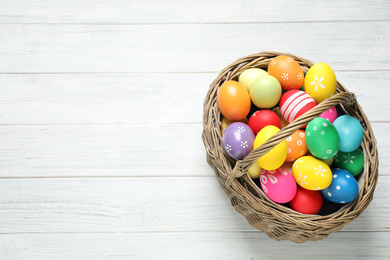 Photo of Colorful Easter eggs in basket on white wooden table, top view. Space for text