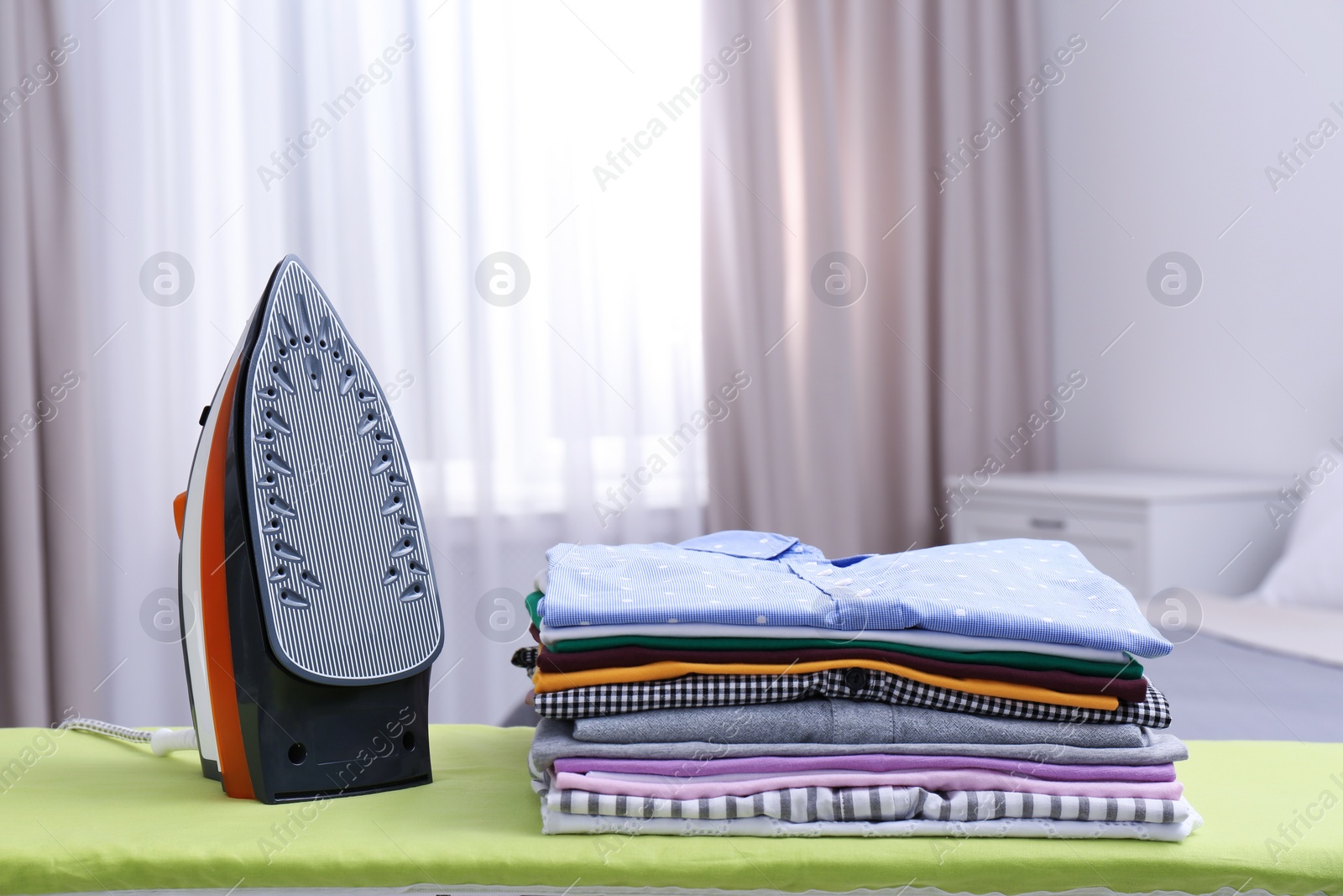 Photo of Modern electric iron and clean folded clothes on board in bedroom. Laundry day