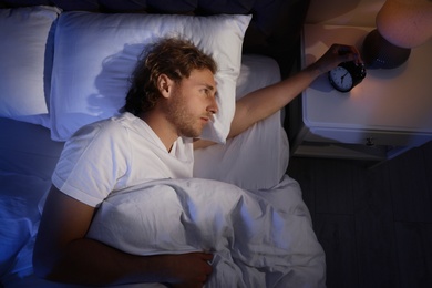 Photo of Sleepy young man turning off alarm clock on nightstand, view from above. Bedtime