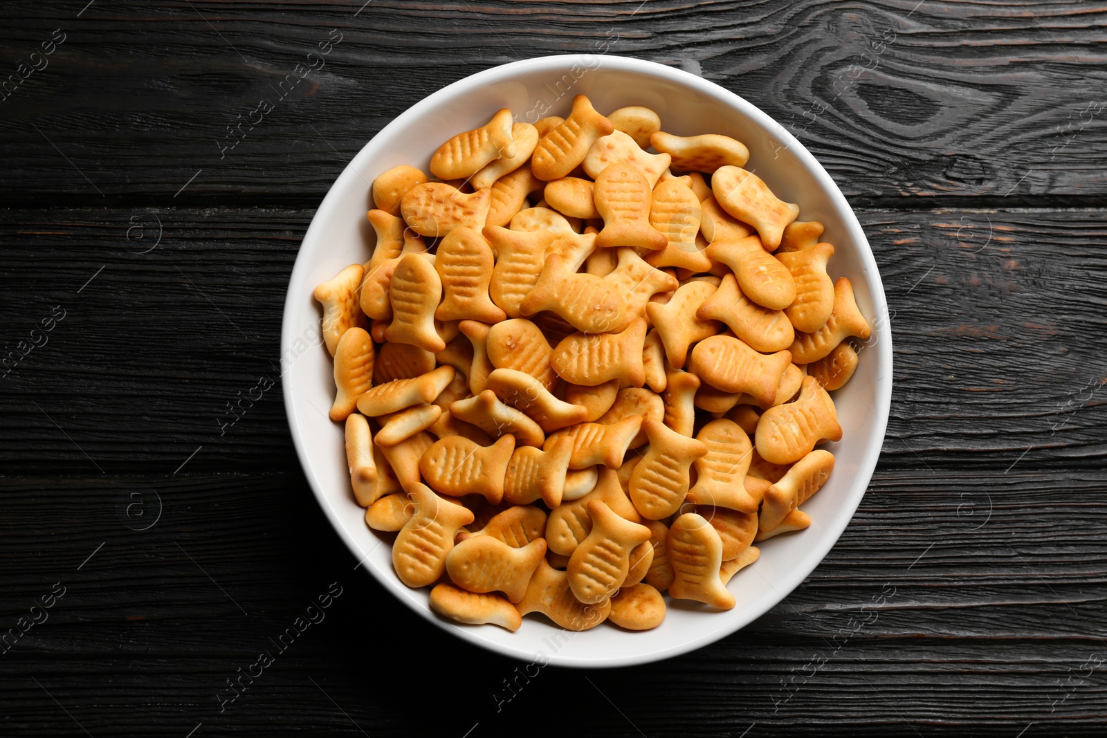 Photo of Delicious goldfish crackers in bowl on black wooden table, top view
