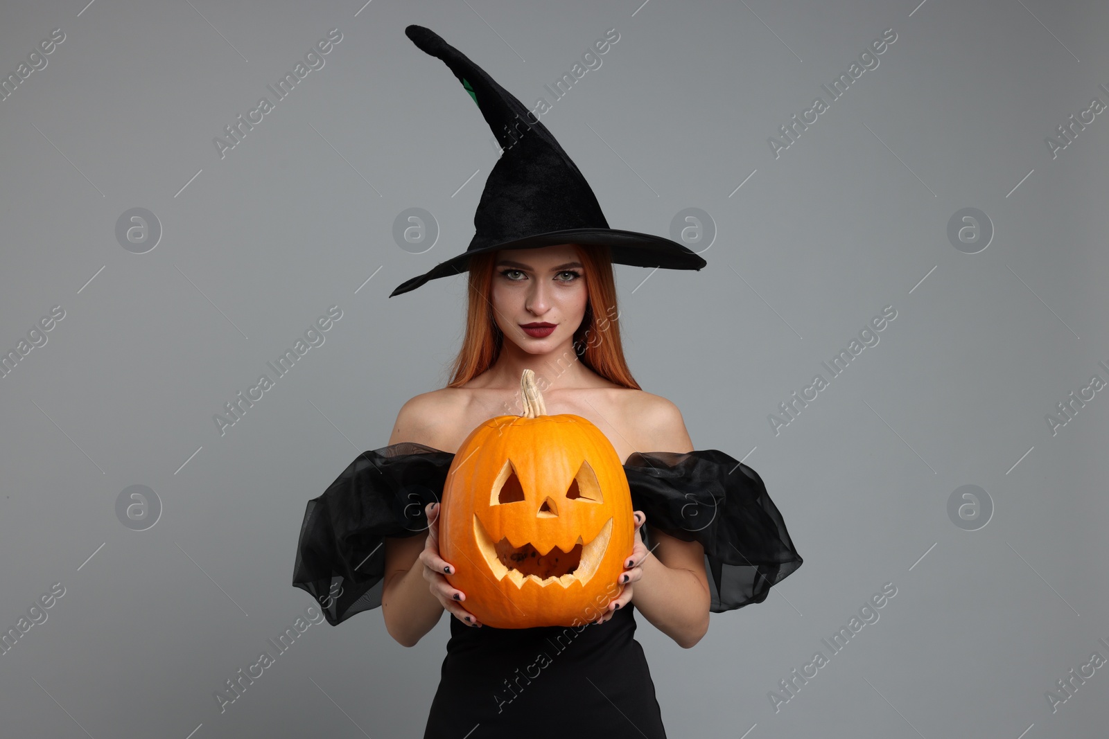 Photo of Young woman in scary witch costume with carved pumpkin on light grey background. Halloween celebration