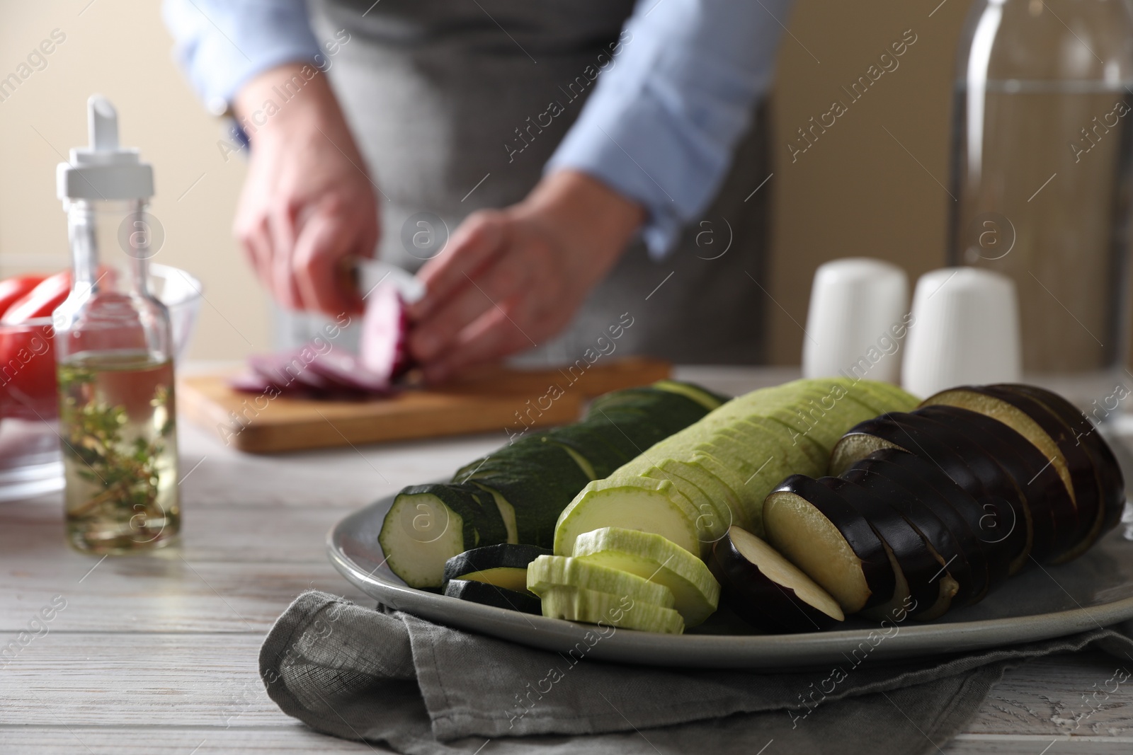 Photo of Cooking delicious ratatouille. Plate with fresh vegetables and woman cutting onion at white wooden table, closeup