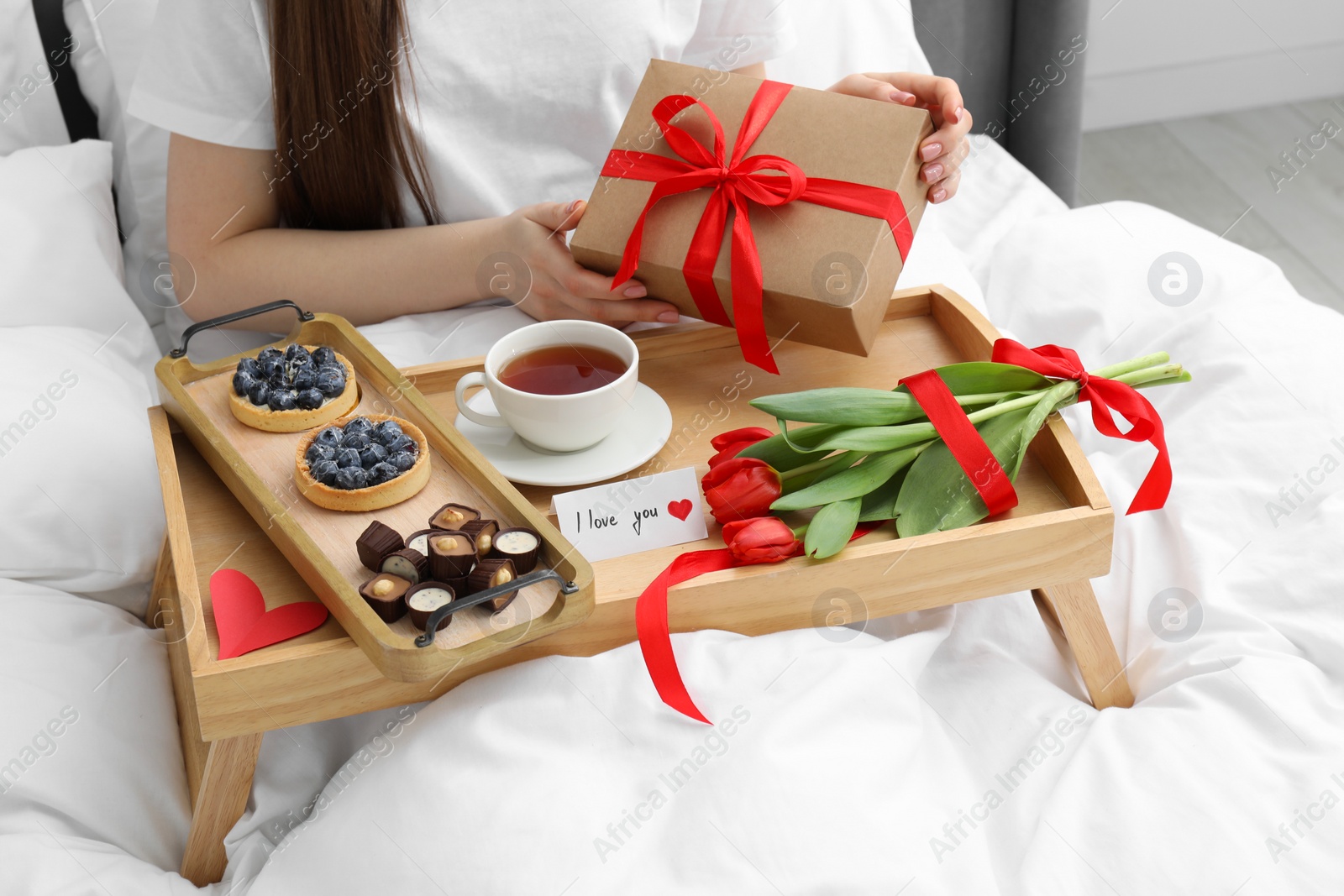 Photo of Tasty breakfast served in bed. Woman with gift box, desserts, tea, flowers and I Love You card at home, closeup