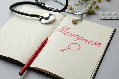 Photo of Stethoscope near notebook with word Menopause and female gender sign on light grey background, closeup
