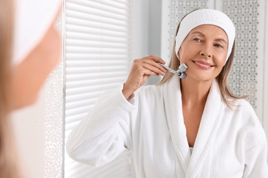 Photo of Woman massaging her face with metal roller near mirror in bathroom