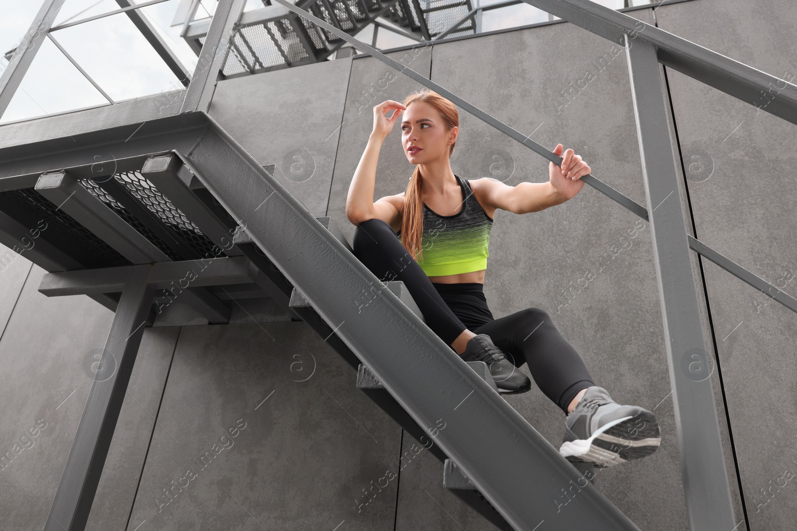 Photo of Beautiful woman in stylish gym clothes sitting on stairs on street, low angle view