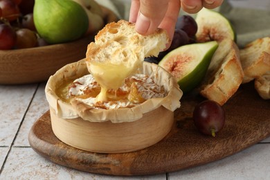 Photo of Woman dipping bread into baked brie cheese at light tiled table, closeup