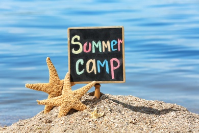 Photo of Little blackboard with text SUMMER CAMP and sea stars on sand near sea