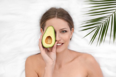 Photo of Beautiful woman with avocado and tropical leaf on white fabric, above view. Organic face mask