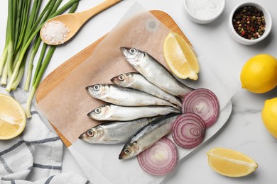 Photo of Fresh raw sprats, cut lemon, onion and spices on white marble table, flat lay