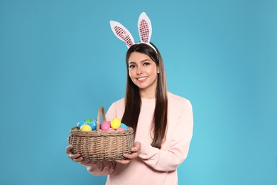 Photo of Beautiful woman in bunny ears headband holding basket with Easter eggs on color background