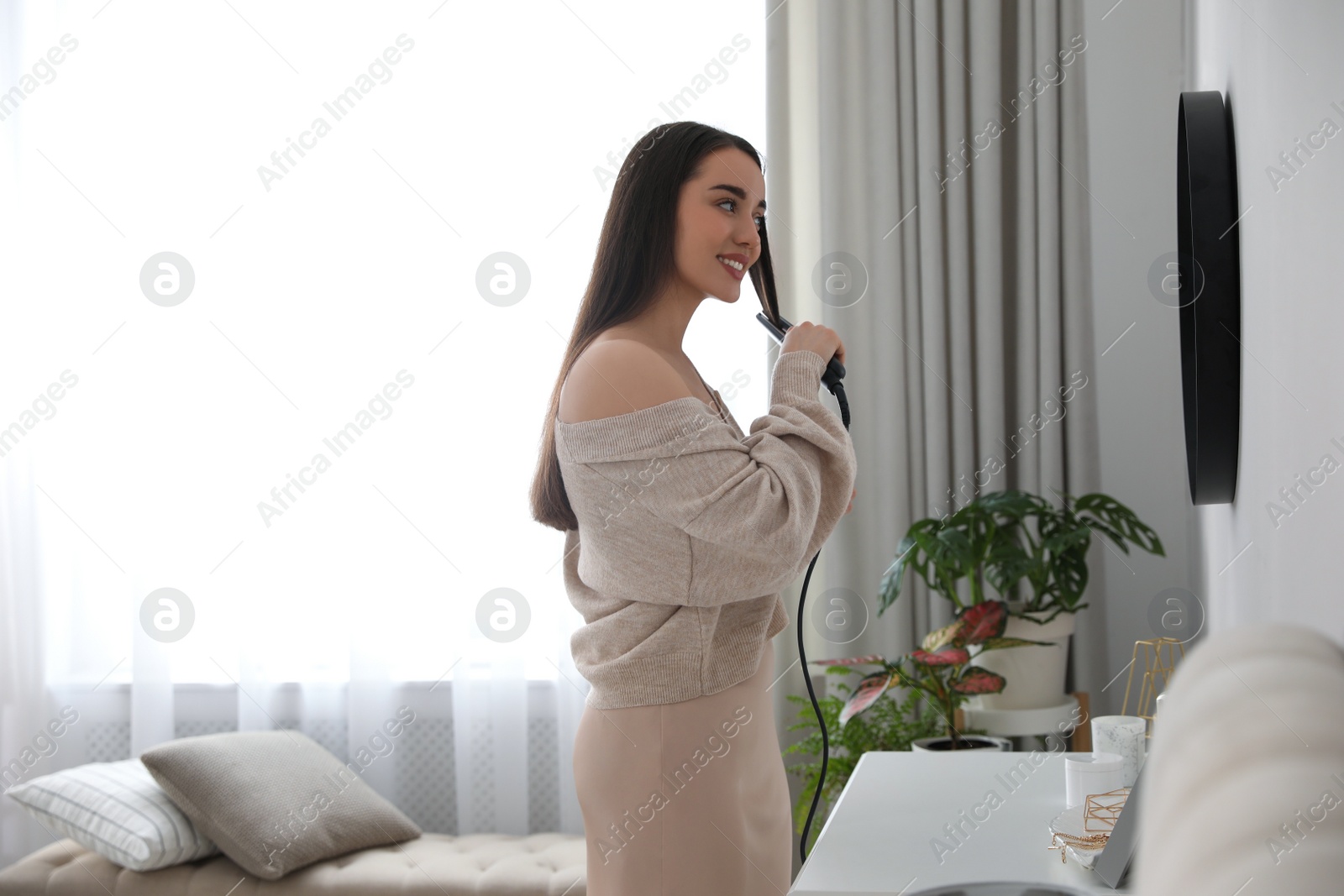 Photo of Young woman straightening hair near mirror at home. Morning routine