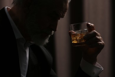 Photo of Senior man holding glass of whiskey with ice cubes on brown background, closeup