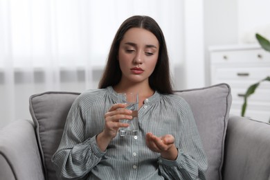 Depressed woman with glass of water taking antidepressant pills in armchair indoors