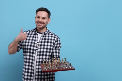 Photo of Smiling man holding chessboard with game pieces and showing thumbs up on light blue background, space for text