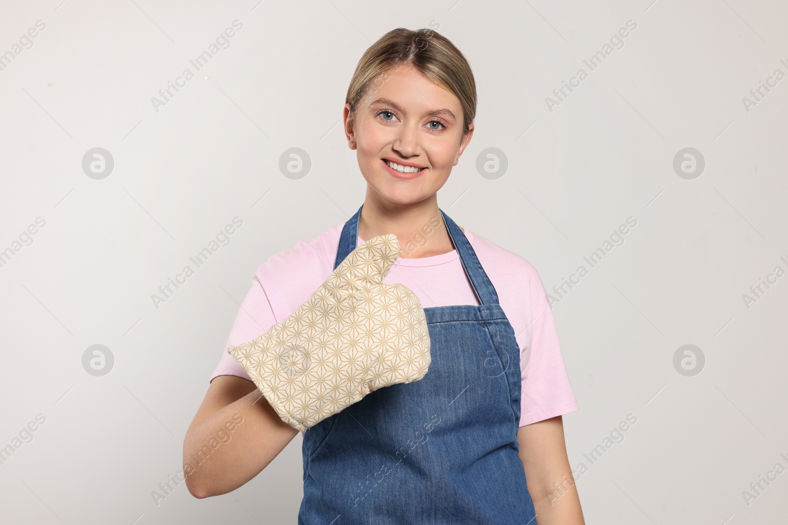 Photo of Beautiful young woman in denim apron and oven glove on light grey background