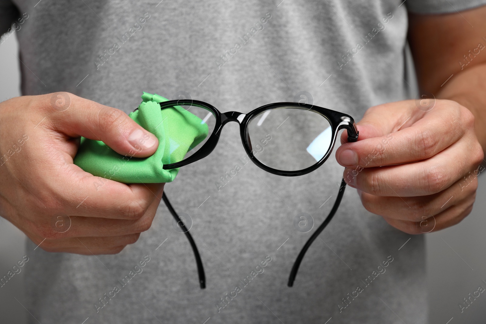 Photo of Man wiping glasses with microfiber cloth on grey background, closeup