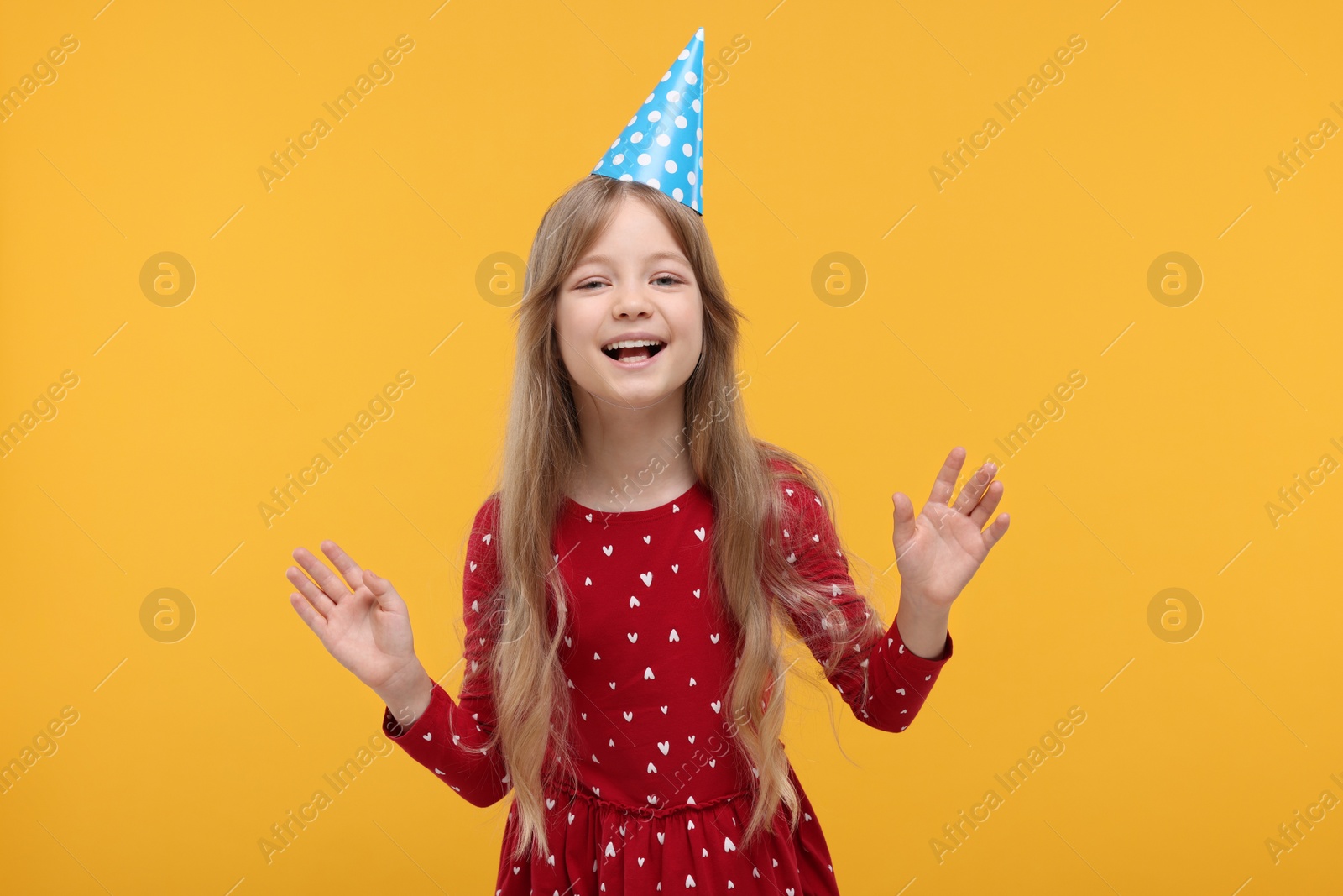 Photo of Happy little girl in party hat dancing on yellow background