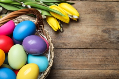 Colorful Easter eggs in wicker basket and tulips on wooden table, flat lay. Space for text