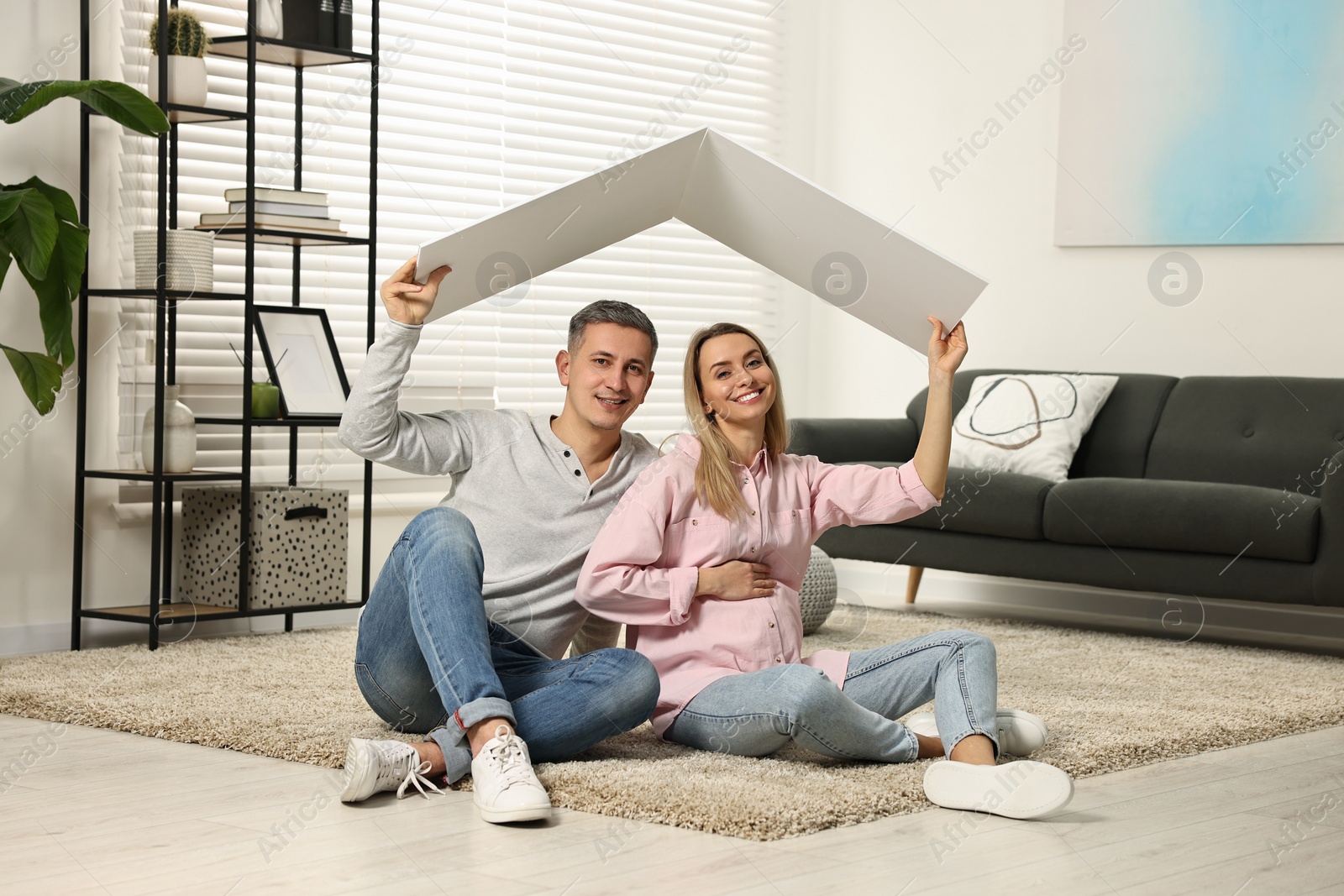 Photo of Young family housing concept. Pregnant woman with her husband sitting under cardboard roof on floor at home