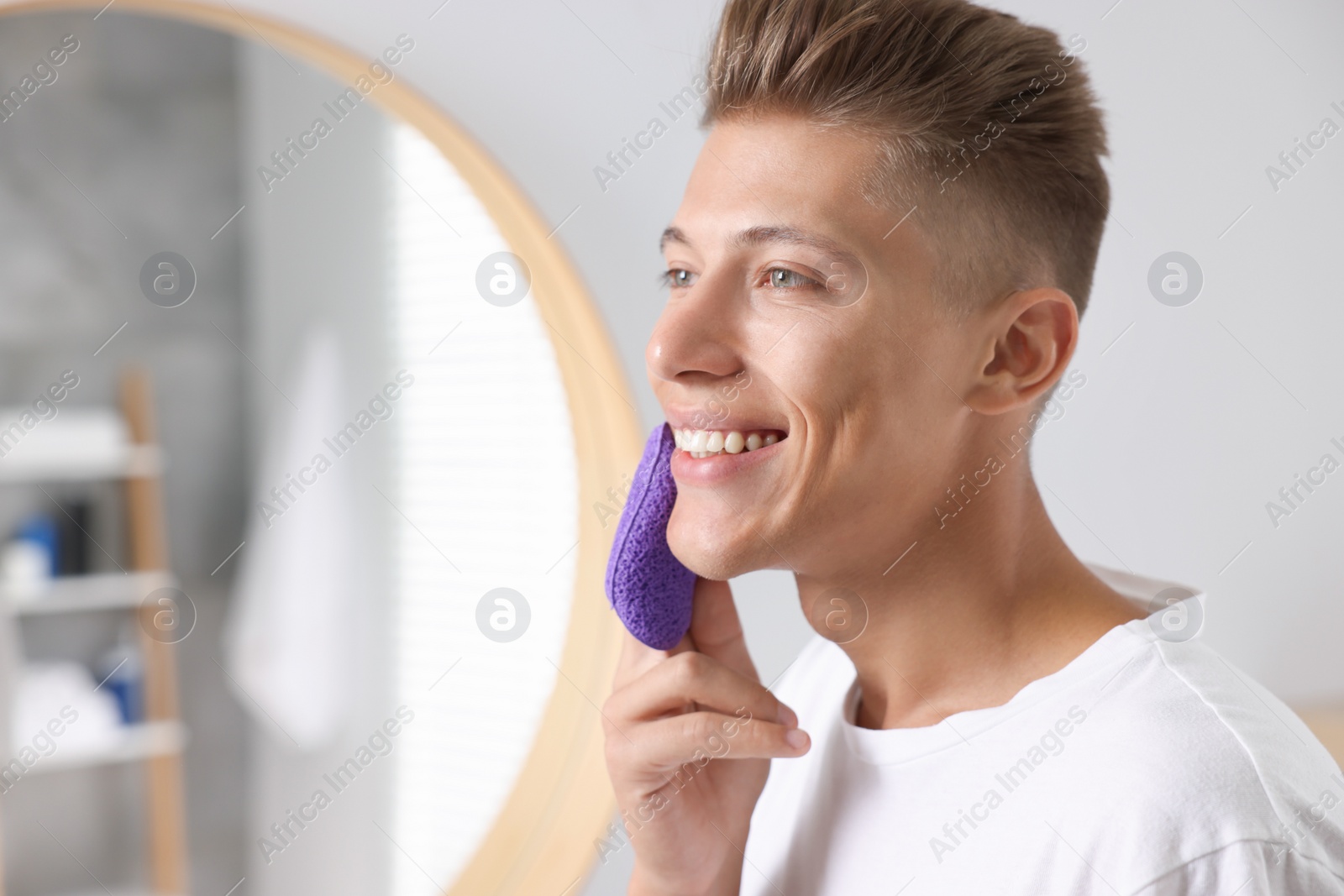 Photo of Happy young man washing his face with sponge in bathroom