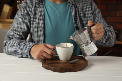 Photo of Man pouring aromatic coffee from moka pot into cup at white wooden table indoors, closeup