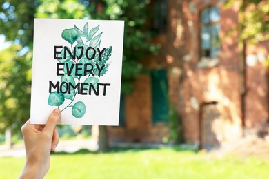 Photo of Woman holding card with phrase Enjoy Every Moment outdoors, closeup. Space for text